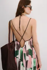 Vestido midi de corte holgado con cuello halter ajustable mediante lazada y hombros descubiertos. Presenta un diseño con tiras cruzadas en la espalda y un estampado abstracto en tonos rosado, verde y marrón sobre fondo blanco.