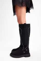 Bota de caña alta confeccionada en cuero vacuno, color negro, con diseño de tachas metálicas dispuestas en forma de arco a lo largo de la caña y correa decorativa con hebilla en la zona del tobillo. Presenta suela track de goma robusta.