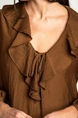 Blusa de color marrón con acabado satinado, cuello en V con volados y lazo de ajuste frontal. Presenta mangas largas con puños elásticos y un corte holgado.