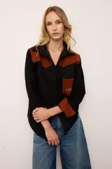 Sobrecamisa de gamuza en color negro con detalles en color marrón en hombros, puños y bolsillo frontal. Presenta bordados decorativos en el bolsillo y un diseño de estilo artesanal.