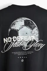 Remera negra de algodón con cuello redondo acanalado y mangas cortas de hombros caídos. Presenta un pequeño estampado tipográfico en el pecho con las palabras "No Defeat Just Glory".