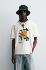 Camiseta de manga corta y cuello redondo, color blanco hueso, con un estampado abstracto multicolor en el pecho que incluye texto en la parte inferior.