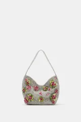 Bolso de hombro plateado cubierto de abalorios y cuentas, decorado con apliques florales tridimensionales en tonos verde y fucsia. Cuenta con un asa corta para el hombro, cierre de clip imantado y bolsillo interior con cremallera.
