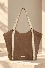 Cartera tipo shopper de gamuza marrón, con correas largas de cuero en tono beige con textura simil cocodrilo y detalles de tachas metálicas plateadas. Cuenta con placa metálica con el logo de la marca en el frente.