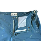 Short de baño color azul petróleo con estampado sutil de palmeras. Presenta una pretina fija con hebillas metálicas laterales para ajuste, cierre frontal con botón a presión, dos bolsillos laterales y un bolsillo trasero. Incluye interior con mesh hipoalergénico.