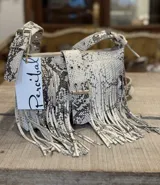 Mini bandolera de cuero con diseño de flecos largos y hebilla frontal decorativa. Presenta un acabado texturizado con efecto craquelado en tonos blanco y marrón, y correa ajustable.