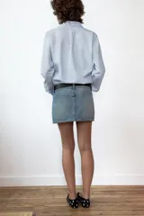 Falda mini de denim en tono azul claro, con tiro medio, trabillas para cinturón y bolsillos de plastrón en el frente. Presenta cierre frontal con cremallera y botones metálicos.