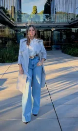 Trench coat gris de corte clásico con doble botonadura, solapas amplias, cinturón ajustable con hebilla y mangas con trabillas en los puños.