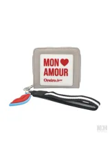 Billetera tipo monedero de material sintético color gris claro, con cierre de cremallera. Presenta un parche frontal blanco con texto en rojo que dice "MON AMOUR" y debajo "Oreiro Love". Incluye un llavero desmontable con forma de labios rojos y una correa de muñeca negra con franjas blancas.