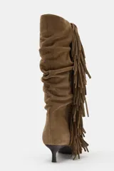 Bota de caña alta confeccionada en serraje, con diseño fruncido y detalle de flecos laterales. Presenta tacón kitten y acabado en punta.