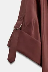 Camisa bordó de cuello alto ajustable con doble hebilla y cierre frontal con botones ocultos por solapa. Tiene mangas cortas con trabillas y diseño holgado.