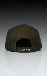 Gorra estilo 5 paneles, color verde militar en la corona y visera plana negra. Presenta un pequeño parche cuadrado negro con un logo blanco bordado en forma de tijeras o lazo.