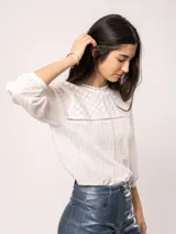 Blusa blanca de algodón con escote redondo, manga tres cuartos y silueta oversized. Presenta bordados y texturas combinadas.