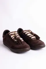 Zapatillas urbanas color marrón, confeccionadas en una combinación de gamuza y tejido texturizado. Presentan un diseño tipo retro con cordones gruesos bicolores en tono crema y marrón, decorados con una borla y un detalle metálico circular en el empeine.