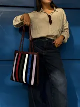 Cartera tipo tote de cuero con diseño de franjas verticales multicolores en tonos tierra, rojo y azul. Cuenta con correas trenzadas de cuero y cierre superior.