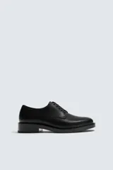 Zapato de vestir tipo derby, color negro, con capellada lisa de cuero y horma en punta. Posee suela de goma con un ligero tacón cuadrado.