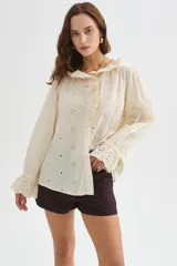 Camisa de algodón con detalles de broderie, volados y cuello a la base. Presenta diseño holgado, cierre frontal con botones y mangas largas con puños fruncidos.