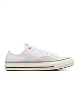 Championes Converse modelo Chuck 70 Ox, de corte bajo, confeccionados en lona blanca con detalles en gamuza gris claro en los laterales. Presentan suela de goma con franjas negras y ojal superior en color rojo.