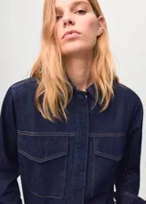 Camisa de denim azul oscuro con cuello camisero, mangas largas con puños abotonados, dos bolsillos delanteros con solapa y cierre frontal de botones ocultos.
