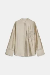 Camisa de cuello mao color beige, confeccionada en tejido satinado con brillo sutil, manga larga con puños anchos y un bolsillo de parche en el pecho con detalles bordados tonales. Cierre frontal con botones ocultos bajo solapa.