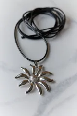 Collar con dije de acero quirúrgico en forma de flor tipo margarita, montado sobre un tiento de cuero negro.