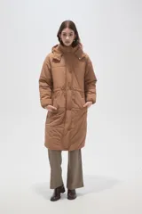 Campera puffer larga de poliamida, color tostado, con capucha desmontable, cierre frontal con botones a presión, bolsillos laterales y cordón ajustable en la cintura.