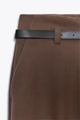 Pantalón de vestir color beige, tiro alto, corte ancho y pierna recta. Presenta un bajo con vuelta y viene acompañado de un cinturón fino de cuero marrón con hebilla metálica.