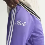 Pantalon deportivo Adidas Originals de corte recto, color violeta con las icónicas tres tiras blancas laterales. Presenta el nombre 'Bob' bordado en la parte frontal y el logo del trébol en la pierna. Cuenta con cintura elástica con cordón de ajuste.