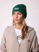 Gorro tipo beanie de tejido acanalado color verde, con parche rectangular frontal con el logo de la marca Rusty en color blanco.