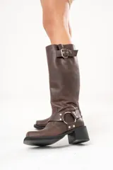 Bota biker de caña alta color marrón, confeccionada en cuero. Presenta hebilla en la parte superior, detalle de aro en la parte inferior y cierre lateral.
