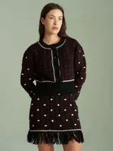 Cardigan de punto negro y rojo con diseño texturizado, cuello redondo y cierre frontal con botones. Presenta detalles de ribetes en blanco y violeta en el cuello, la botontera, los puños y el ruedo, que cuenta con flecos colgantes.