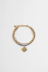 Pulsera doble de acero dorado, compuesta por una cadena de eslabones largos y otra con cuentas metálicas y cuentas de piedra azul. Cuenta con un dije central redondo con diseño de ojo turco y cadena de alargue con terminal en forma de corazón.