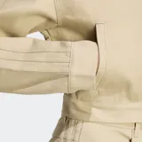 Campera Adidas Firebird color beige, con cierre frontal y cuello alto. Presenta las tres tiras características de la marca en las mangas y logo bordado en el pecho. Corte por encima de la cintura.