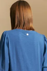Camisa de algodón blanca con delicadas líneas bordadas verticales y textura calada. Presenta escote redondo con cierre frontal de botones, mangas largas con leve volumen en los hombros y puños elásticos.