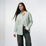 Camisa de corte oversize en color verde con estampado de rayas verticales finas. Presenta cuello clásico y puños en color blanco, cierre frontal con botones y un bolsillo aplicado en el pecho.