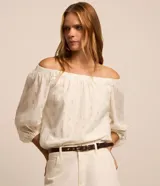 Blusa de tricolina blanca con estampado de lunares dorados en foil, escote descubierto estilo hombro a hombro y mangas largas abullonadas con puños elásticos.