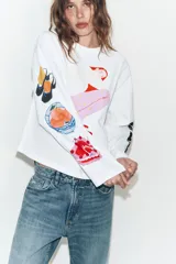 Camiseta blanca de corte oversize con cuello redondo y manga larga. Presenta una estampa gráfica central de una figura femenina, detalles estampados en la manga derecha y texto en la manga izquierda.