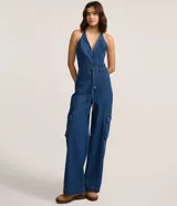 Mono largo de jean azul, sin mangas, con escote en V y cuello halter. Cuenta con cinturón de lazo en la cintura y bolsillos cargo en los laterales de las piernas.