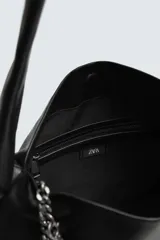 Bolso tipo bandolera de cuero sintético negro, con asa de hombro y detalle de cadena metálica plateada en el frente. Incluye un charm colgante con forma de llave.