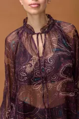 Blusa de gasa translúcida con estampado paisley en tonos burdeos, beige y azul. Presenta cuello alto con volante fruncido, abertura frontal con cordones ajustables y mangas largas abullonadas con puños elásticos.