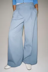 Pantalón wide leg de tiro medio confeccionado en hilatura de algodón, color beige, con trabillas en la cintura, bolsillos laterales ocultos y detalle de pliegues en el delantero. Cierre frontal con cremallera, botón y ganchos metálicos.
