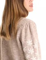 Buzo de punto marca Pinch, color marrón claro, con cuello redondo y mangas largas. Presenta un diseño de flores blancas bordadas a lo largo de las mangas y detalle de frunce en los hombros. Cuenta con puños y ruedo acanalados.