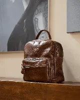 Mochila de cuero natural color marrón con acabado texturizado. Presenta un compartimento principal con cierre, un bolsillo frontal con cierre y asa superior. Diseño clásico y funcional con costuras reforzadas.