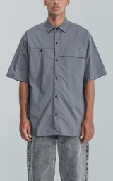 Camisa de manga corta en color gris, confeccionada en popelina de algodón. Presenta un corte oversize, cuello clásico, cierre frontal con botones y dos bolsillos delanteros, uno de ellos con solapa.