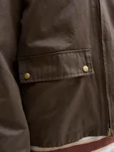 Campera de estilo worker color marrón, con cuello camisero de pana, cierre frontal con cremallera y bolsillos frontales con solapa y broches a presión. Presenta un acabado recubierto que le otorga una textura sutilmente brillante y resistente.