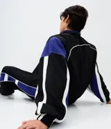 Campera deportiva de poliamida con diseño de bloques de color en negro, blanco y violeta. Presenta cuello alto, cierre frontal con cremallera, puños y ruedo elásticos, y forro interior de poliéster.