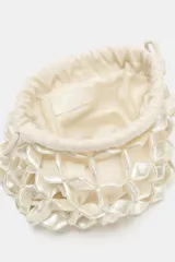 Bolso pequeño tipo saco de mano, con cuerpo exterior compuesto por cuentas facetadas nacaradas que forman un patrón de red. Posee un forro interior de tela color crema con cierre fruncido y cordones laterales. El asa de mano está formada por las mismas cuentas nacaradas.