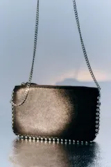 Cartera pequeña color dorado metálico con cierre superior. Presenta tachas plateadas decorativas a lo largo de los bordes superior e inferior.