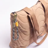 Cartera tipo tote de color khaki, con diseño acolchado de ondas y doble asa de hombro. Incluye un detalle de cordón decorativo con mosquetón en el lateral y una placa metálica pequeña en la parte inferior frontal.