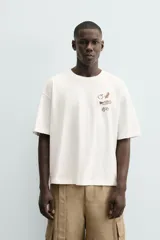 Camiseta de manga corta color blanco roto, corte relajado, con un pequeño bordado/estampado en el pecho que incluye una manzana, una rama de laurel, una trompeta y dos dados.